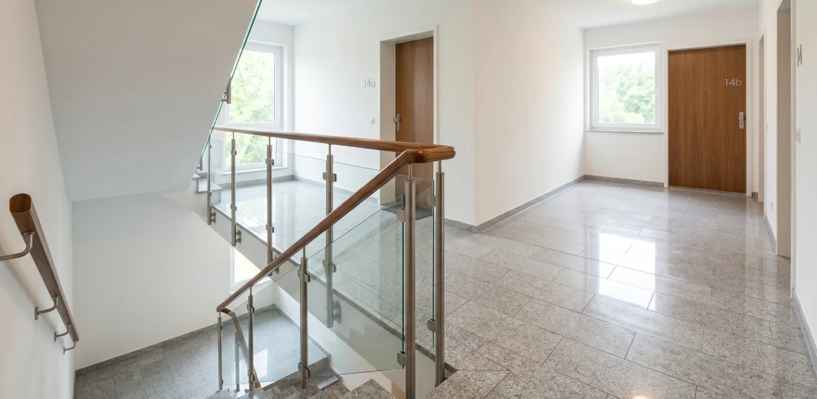 Treppenhaus-Etage nach der wöchentlichen Reinigung: glänzender Granitboden, sauberes Glasgeländer mit Holzhandlauf und freie Wohnungstüren.