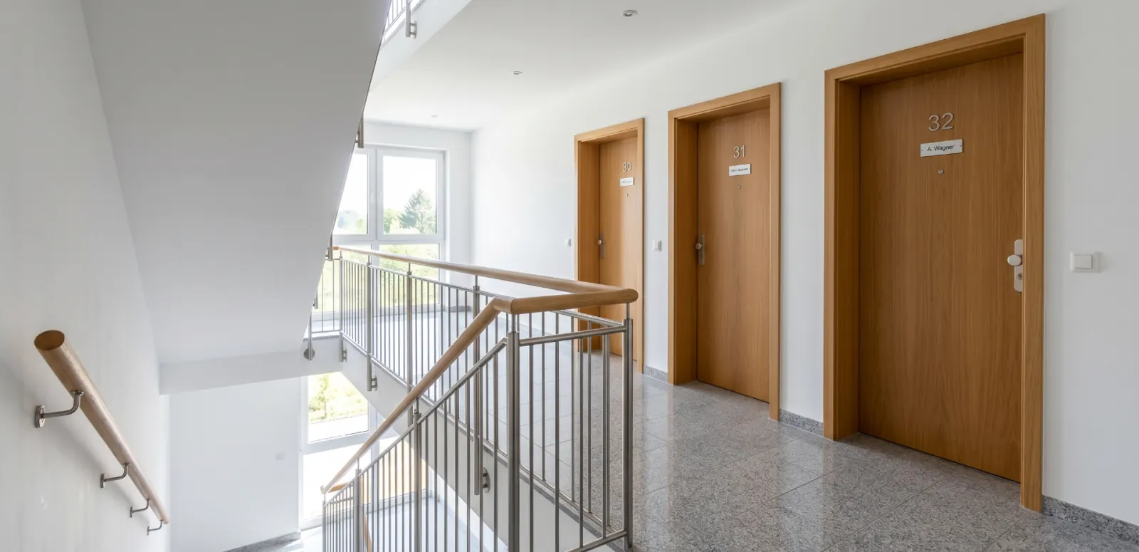 Obere Etage eines Mehrfamilienhauses nach der wöchentlichen Treppenhausreinigung: glänzender Granitboden, sauberes Edelstahl-Glasgeländer mit Holzhandlauf, drei freie Wohnungstüren mit polierten Türschildern.