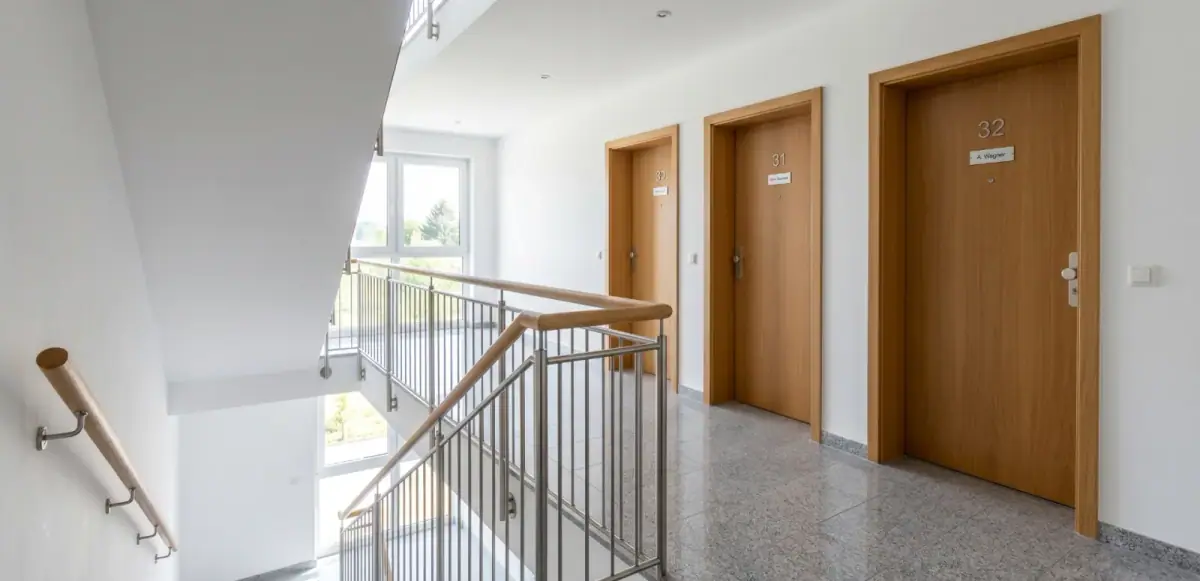 Obere Etage eines Mehrfamilienhauses in Bonn nach der wöchentlichen Treppenhausreinigung: glänzender Granitboden, sauberes Edelstahl-Glasgeländer mit Holzhandlauf, drei freie Wohnungstüren mit polierten Türschildern.