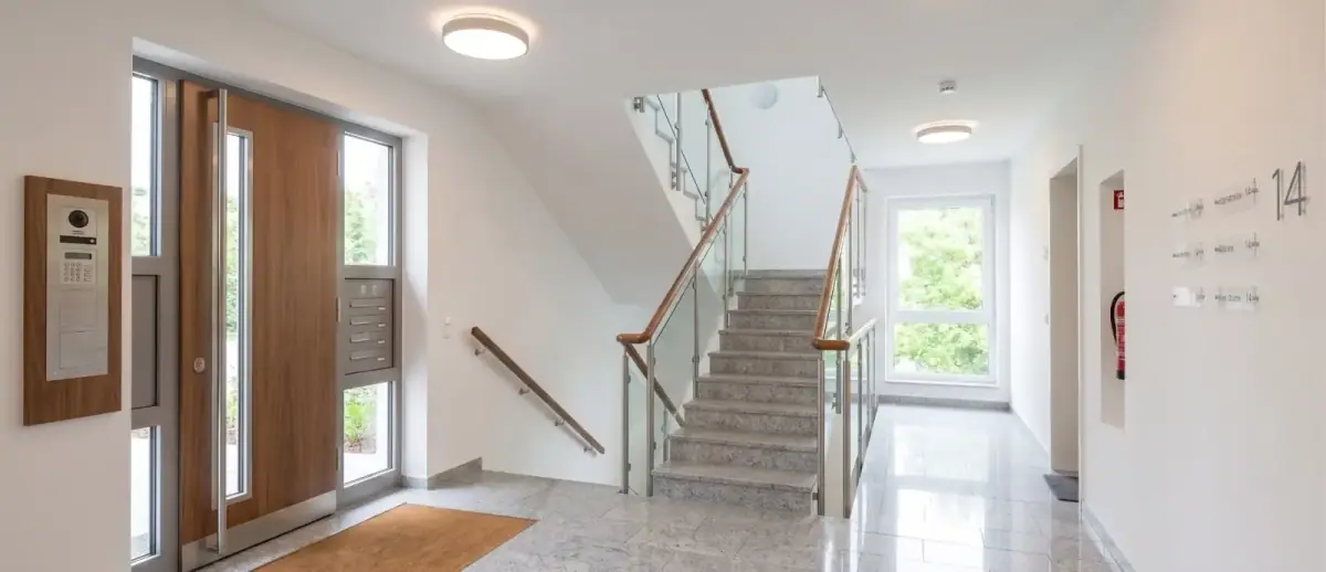 Heller Eingangsbereich eines Mehrfamilienhauses in Köln nach der Treppenhausreinigung: gewischter Fliesenboden mit Spiegelung, saubere Glas-Eingangstür mit Tageslicht und ordentlich abgelegte Willkommen-Matte.