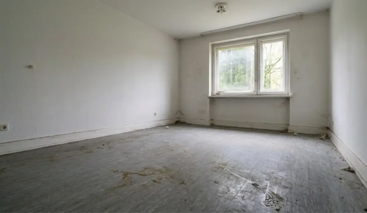 Leerer Raum vor der Grundreinigung: matter, fleckiger Boden, staubige Fensterbänke, sichtbare Spuren auf Sockelleisten, fahles Licht.